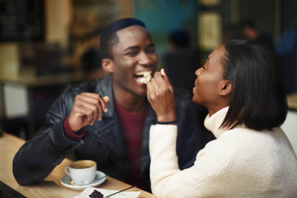 A woman feeds a man food on a date while laughing.
