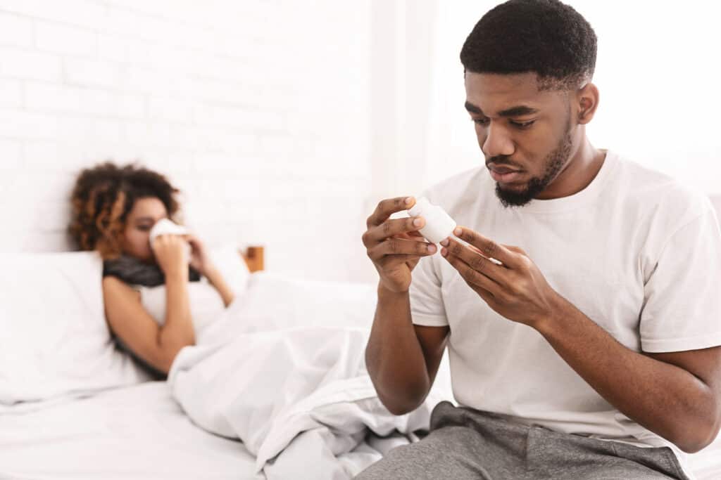 A man is reading medication while a woman sniffles sickly in the background.