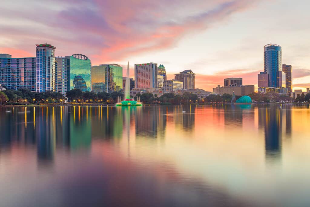 A city skyline with a lake in front under a sunset sky.