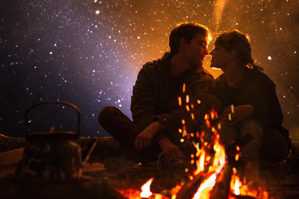 A couple sits outside near a fire under a starry sky.