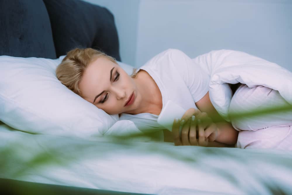 A woman lays in bed looking anxious while on her phone.