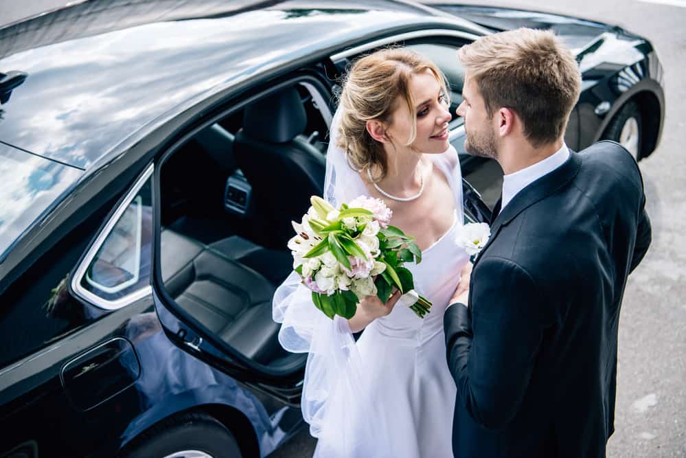 A man and woman in wedding clothing lean in for a kiss by a car.