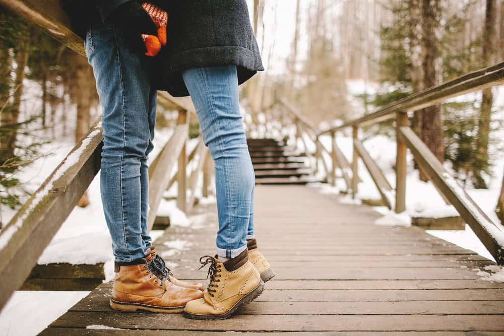 A couple\'s feet outdoors on a bridge covered in snow. The woman is on her tip toes.