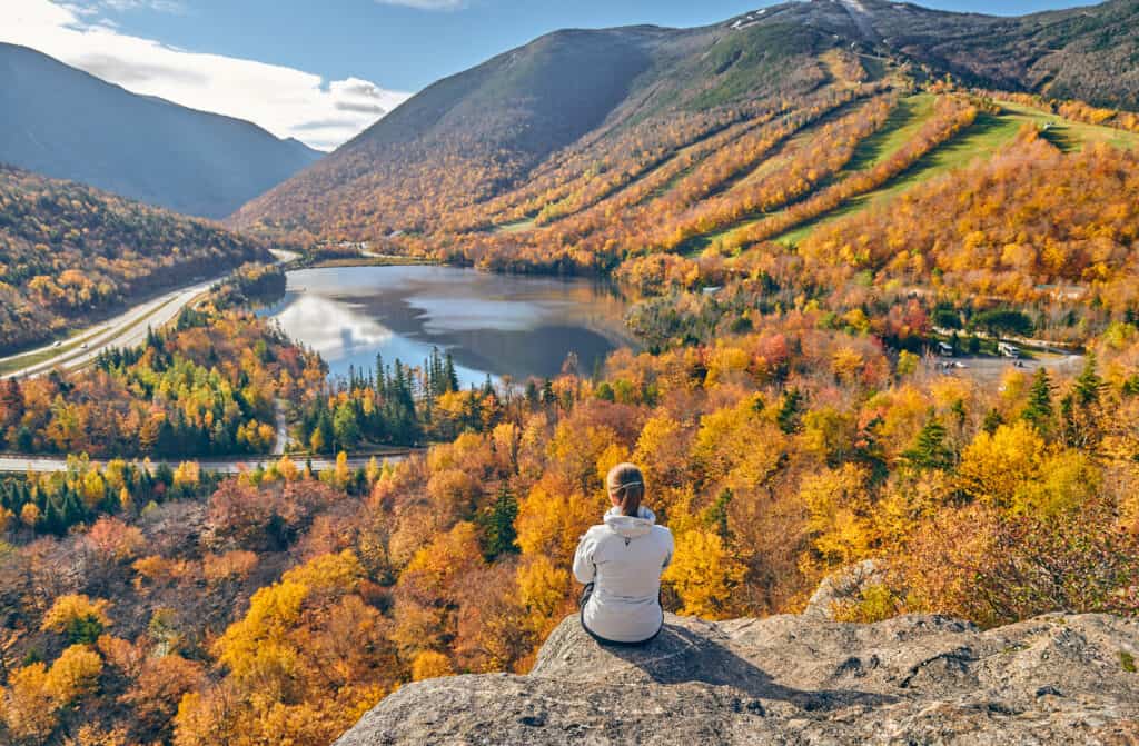 A woman sits on a mountain overlooking a lake with fall-colored mountains.