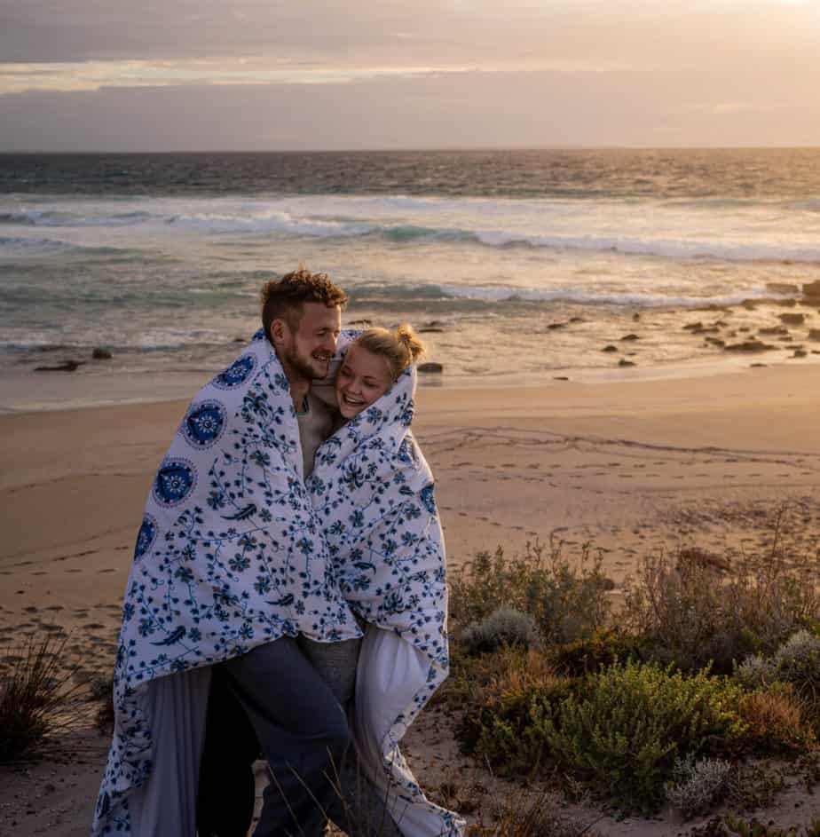 A couple snuggles in a blanket while standing at a beach at sunset.