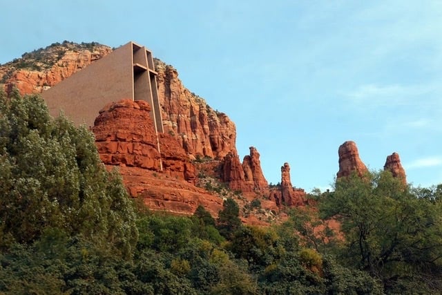 A large building on the side of a red rock formation with green trees along the ground below it. 