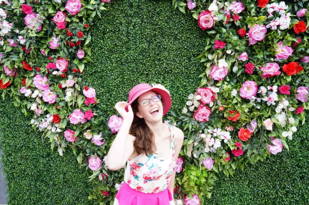 A girl stands while wearing pink in front of a wall filled with pink flowers.