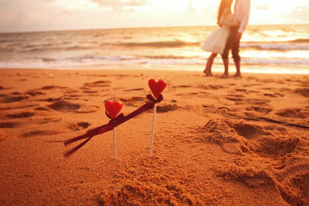 A couple walks on the beach in the background. In the foreground, two sticks with hearts stick out of the sand.