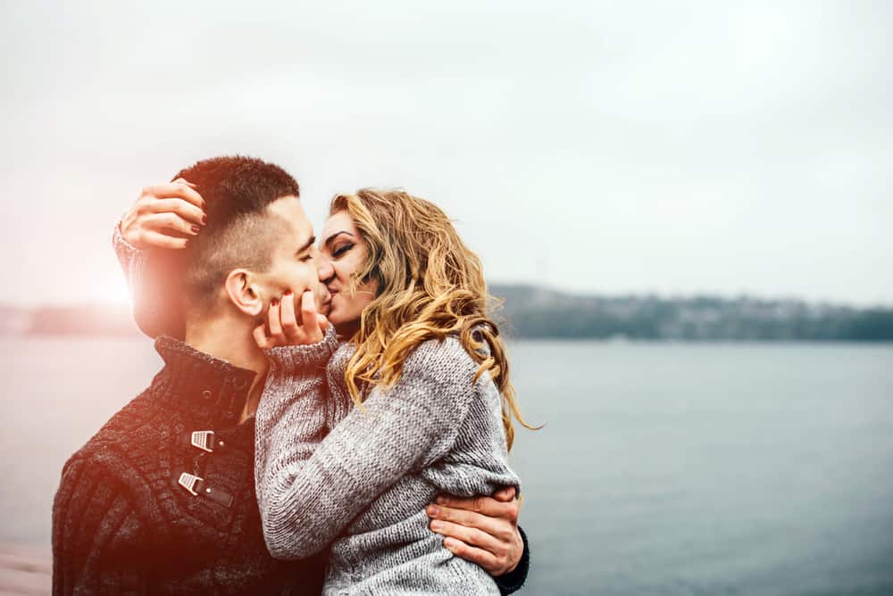 best date ideas for couples header image - brown haired man holding blond woman like a baby in front of a lake on a gray day, they are kissing