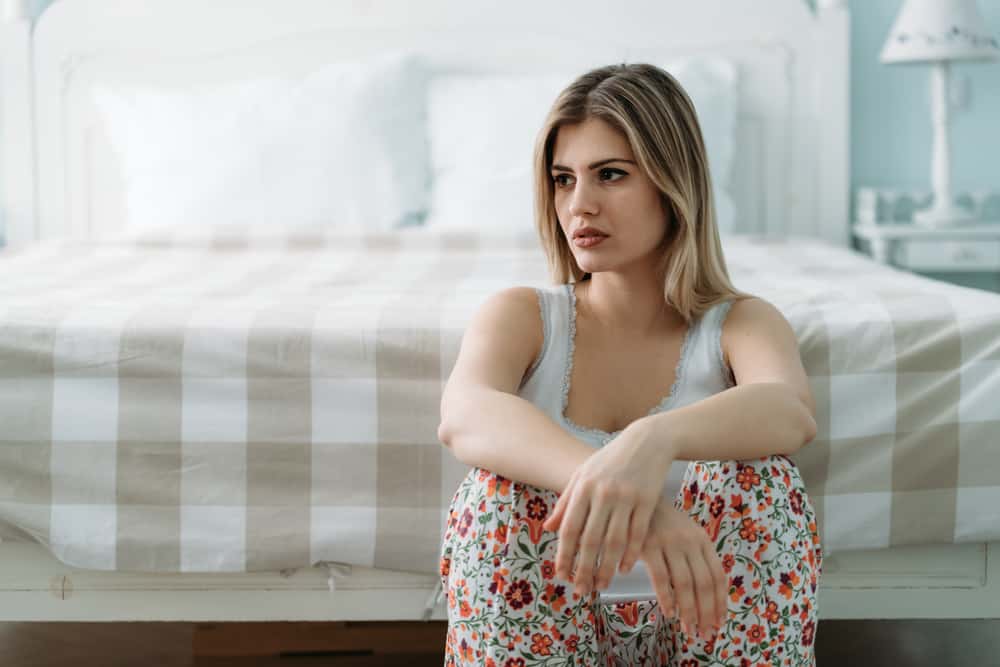 A woman sits upset on the floor next to her bed.