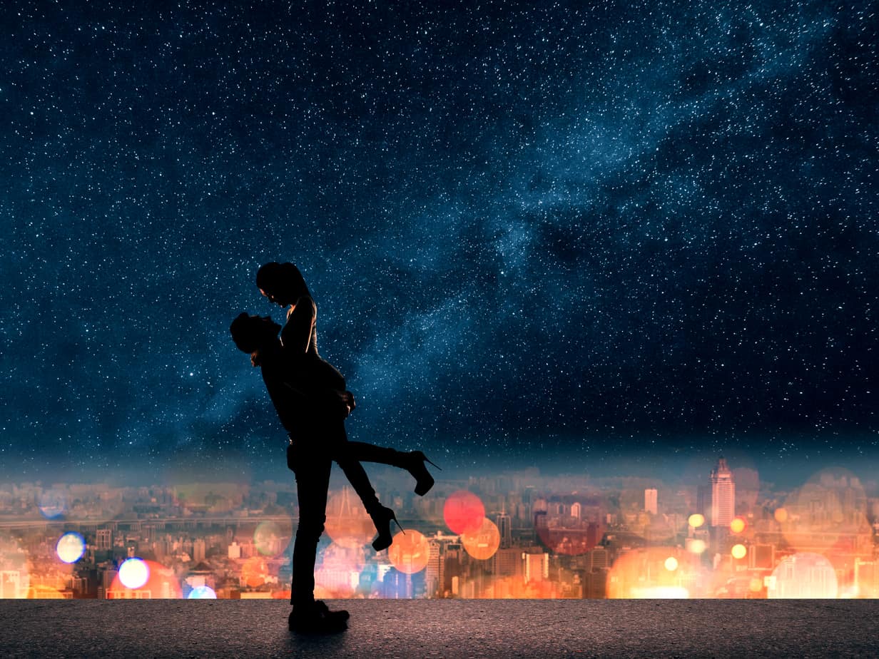 Silhouette of a couple under the stars. A man is picking up a woman in heels with a city skyline behind them.