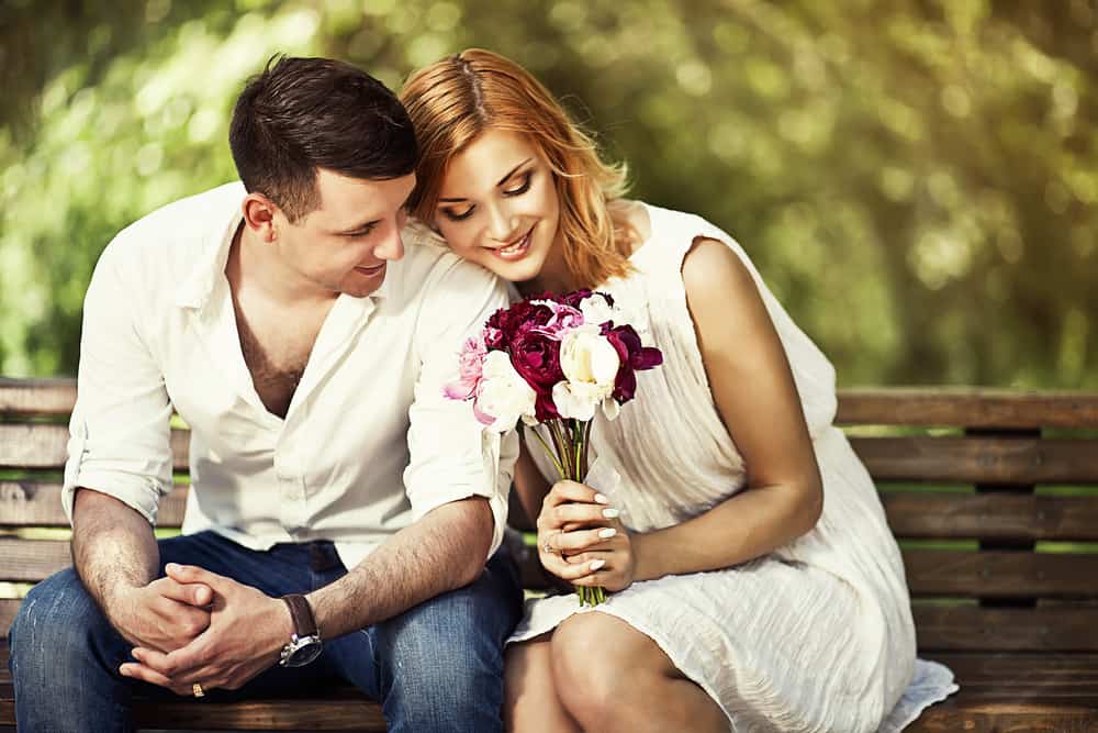 A couple sits on a park bench while the woman holds flowers.