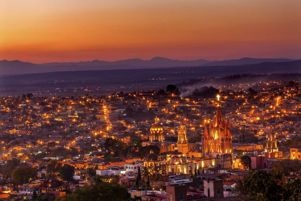 View of a city from above with mountains in the background under a pink and orange sky.