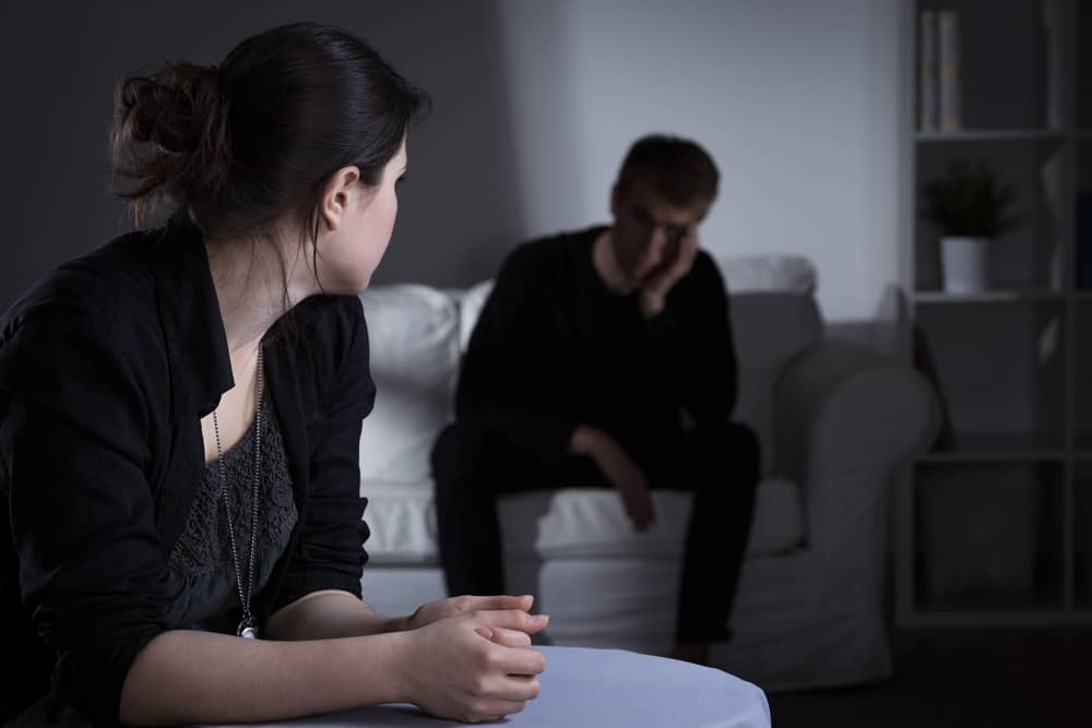 A woman sits while looking back at a man who is upset sitting on a couch.