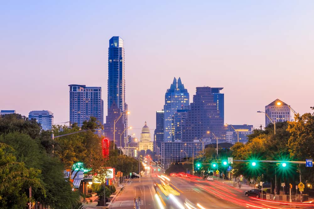 Cars rush past in a city with tall buildings under a purple and pink sky.