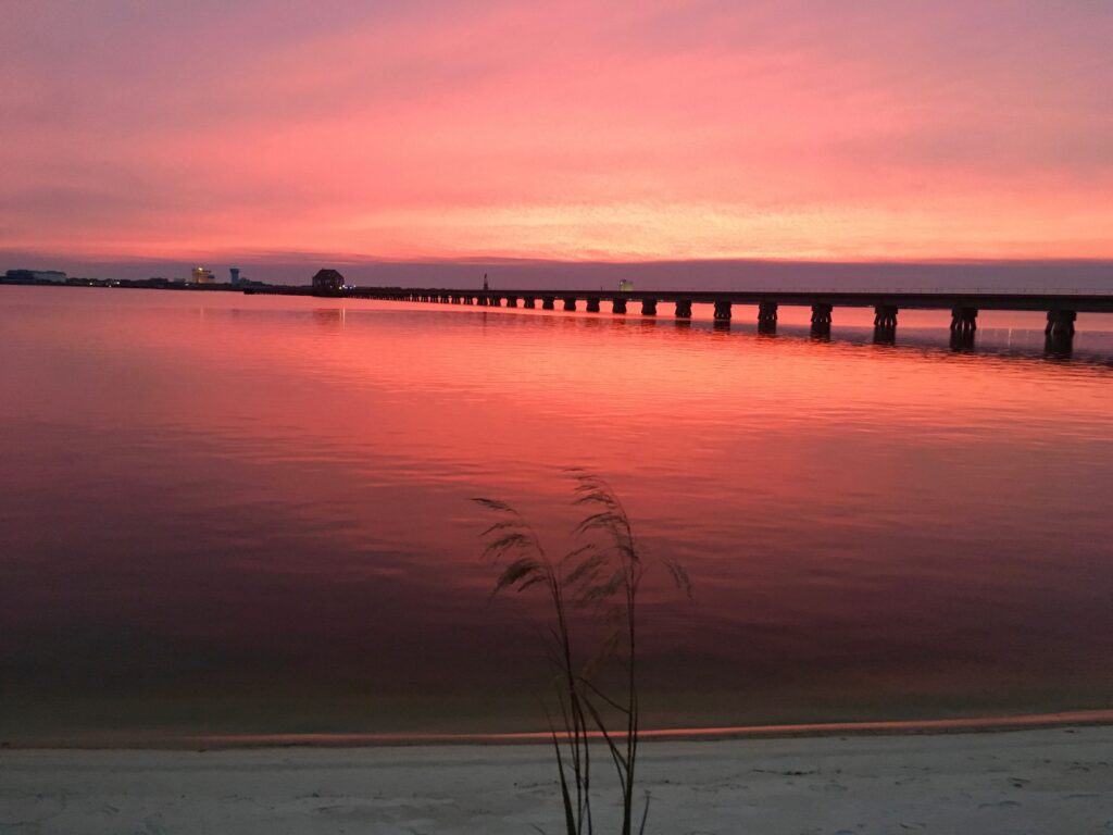A long pier stretches over the ocean at sunset, creating a pink hue.