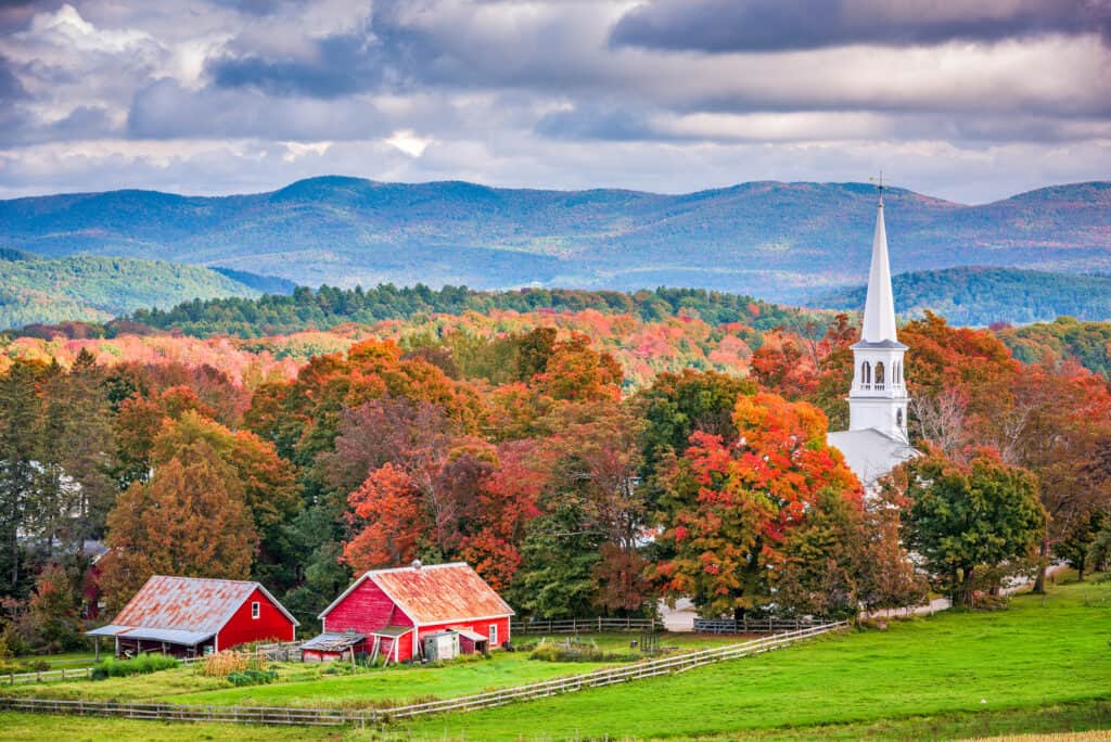 fall is the perfect time for romantic getaways in vermont - image of fall foliage covered mountains behind red barn and white church steeple, classic pastoral shot