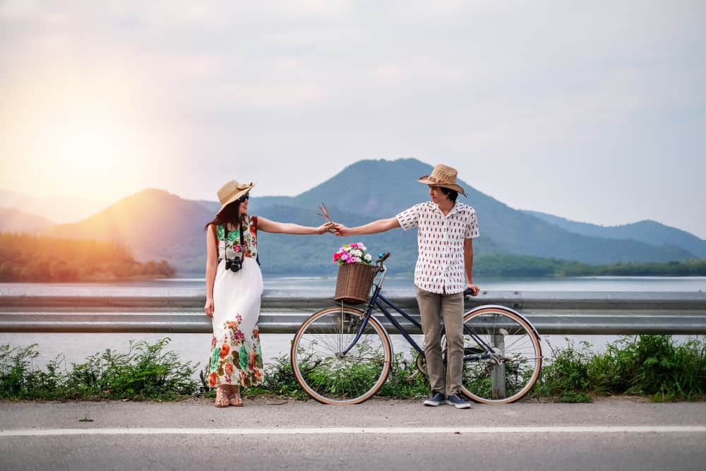 A couple touches hands in front of a sunset and a bike with water and mountains behind them.