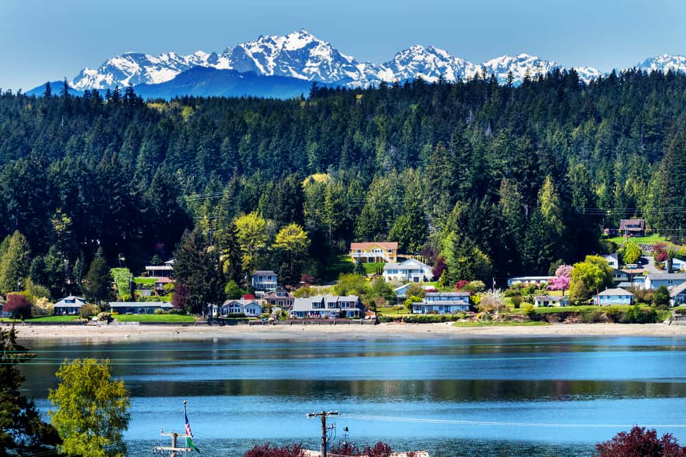 A calm lake sits with a set of buildings behind it. A forest and snow-capped mountains are in the distance.