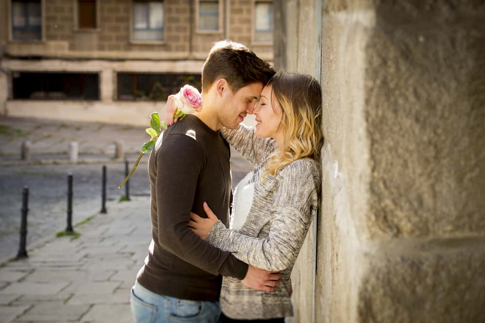 A couple embraces against a wall outdoors while the woman holds a flower.