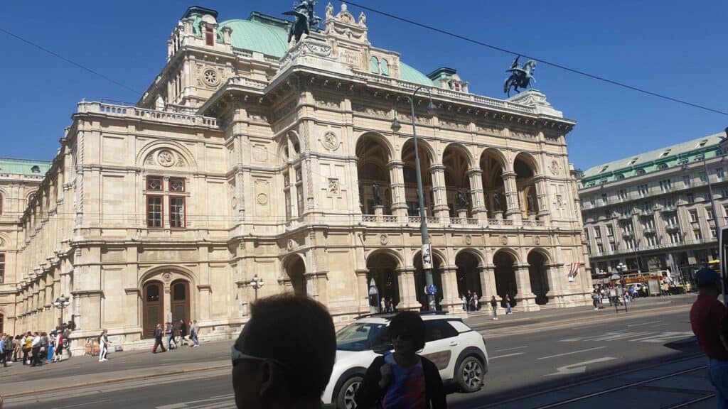 Cars and people rush by with a big building with statues on top behind.