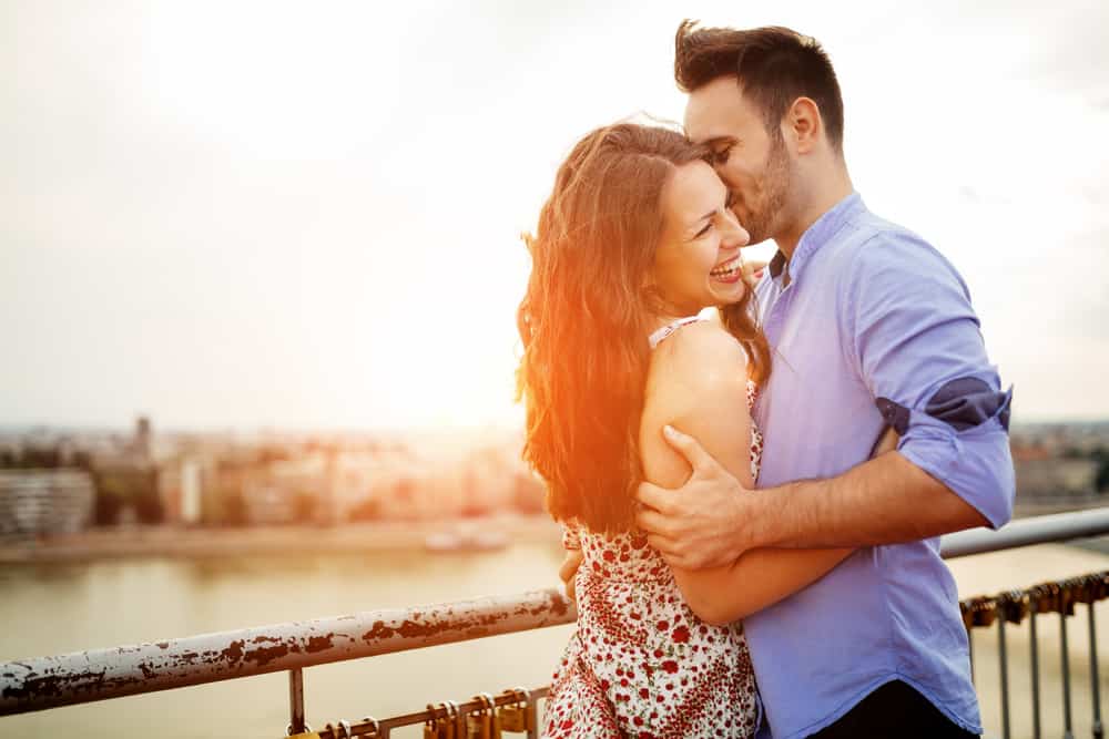 Couple in love hugging outdoors, woman is laughing as man whispers in her ear. The sun peeks out behind them.