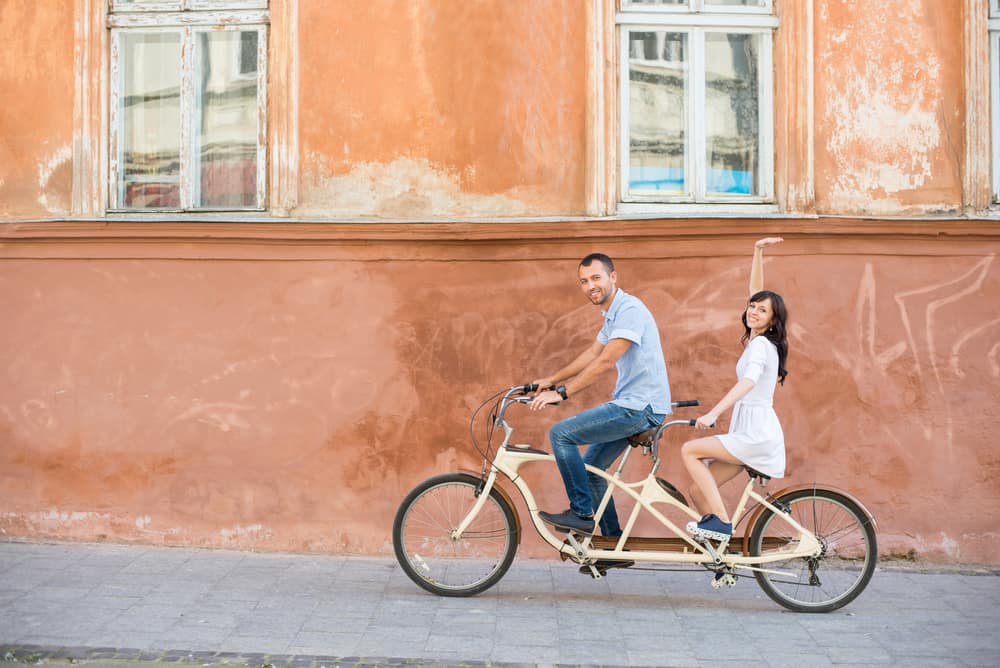 A happy couple rides on a retro tandem bike on the street against the background of an old orange wall with windows. The man is at the front of the bike while a girl in a white dress is raising her hands up in the back, excited.