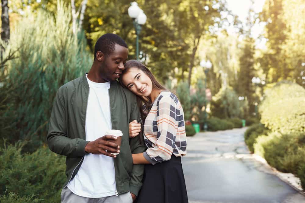 A woman cuddles a man on an outdoor walk, while he holds a cup of coffee. They both look happy.