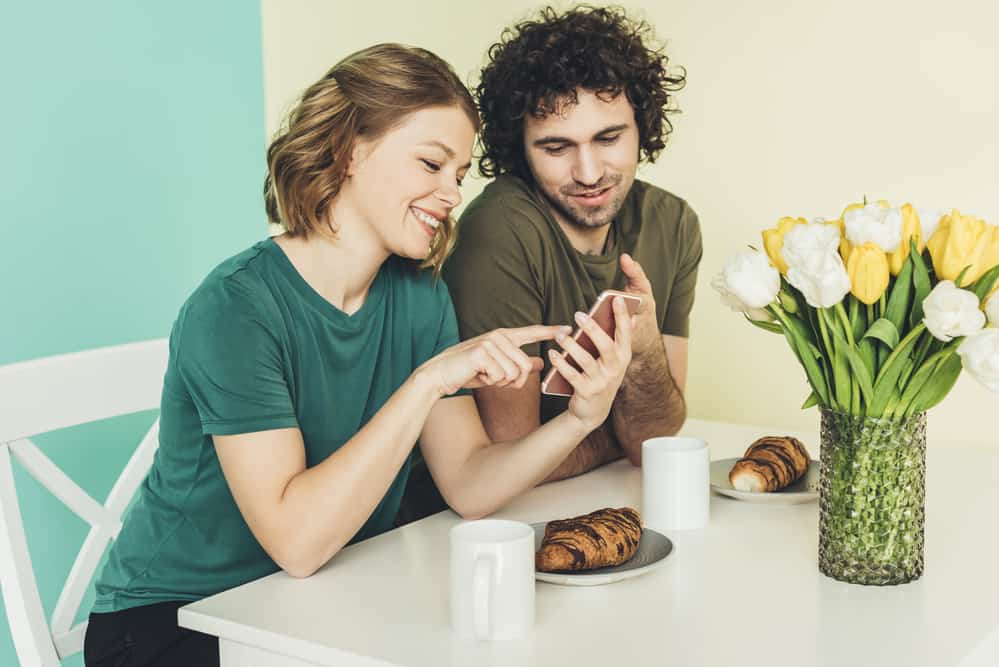 A man and woman sit at a table with croissants and mugs. The woman points on a phone and the man looks.
