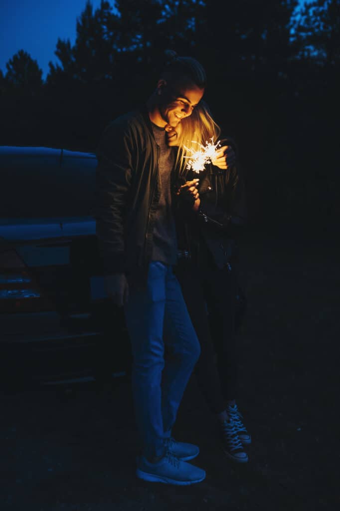 A couple cuddles in front of a car while holding a sparkler.