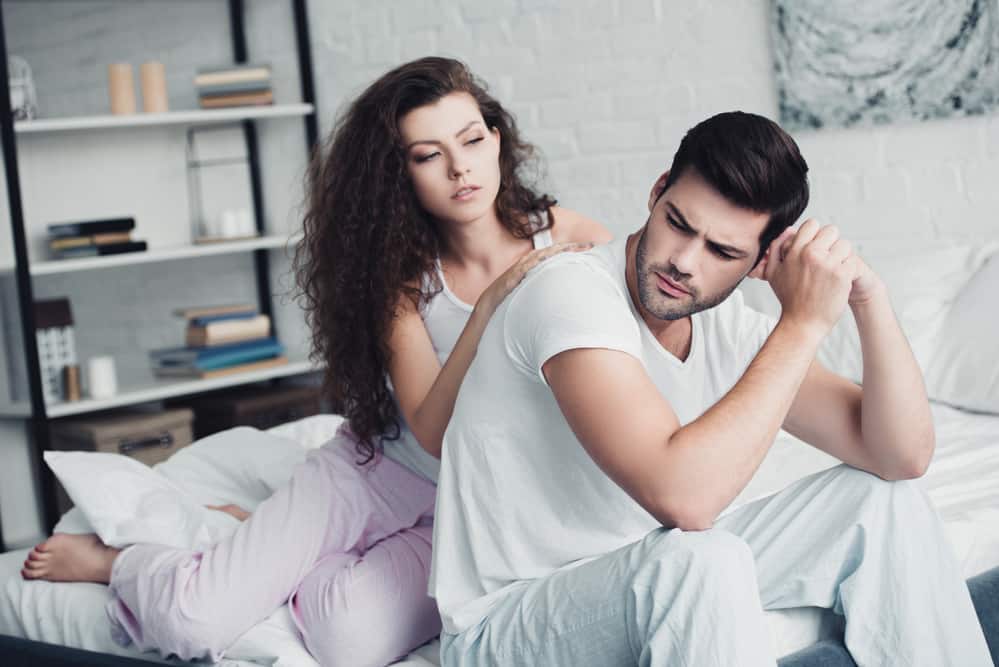 A woman leans on a man\'s back while sitting on a white bed.