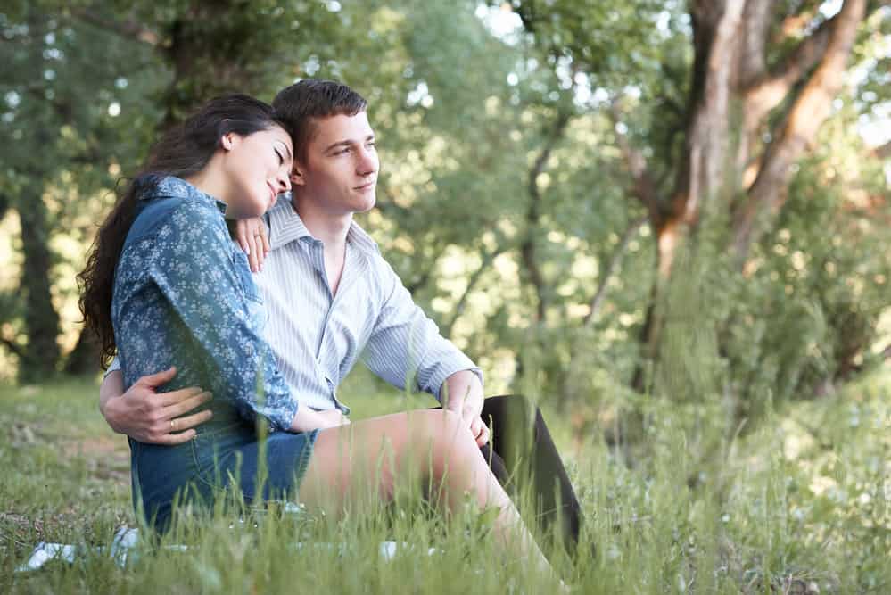 A couple sits on the grass in a forest and looks out into the distance.