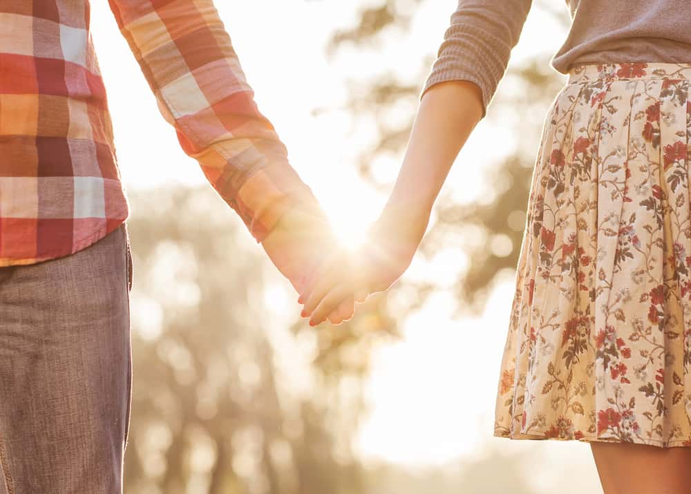 A couple holds hands as the sun peeks through the trees behind them.