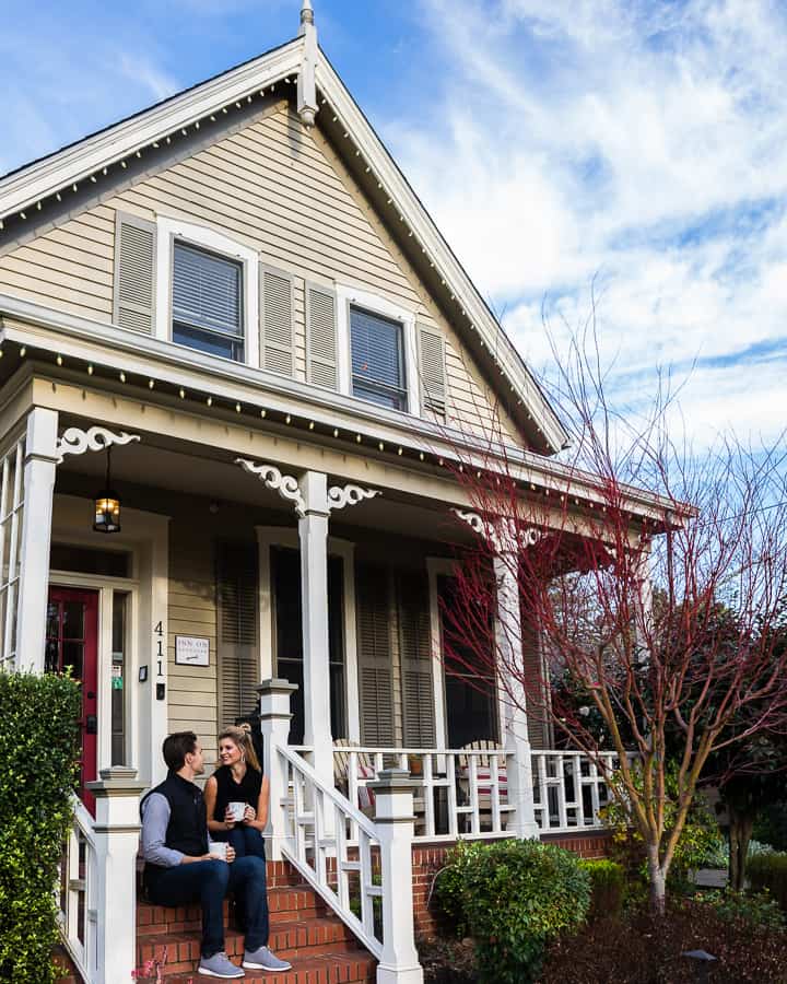 A couple sits on a brick stairwell in front of the house while holding white mugs.