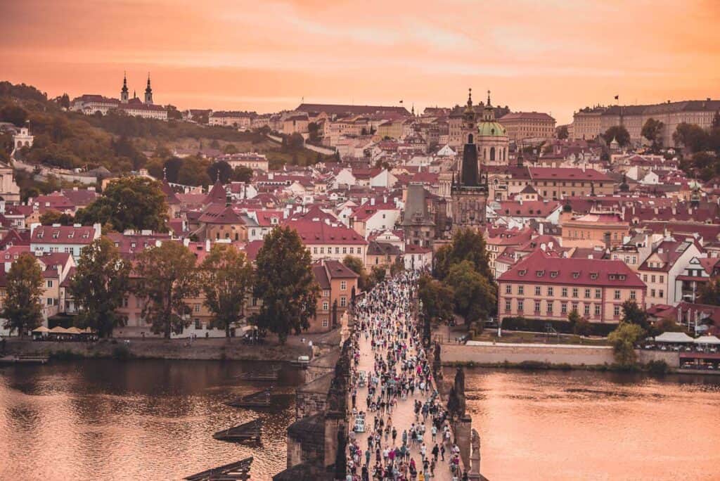 A crowd of people walking across a bridge over a river leading to a European town. 