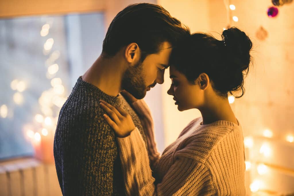 Passionate romantic couple in sweaters are spending time together before New Year at home. Tender couple hugging on the background of shiny garland.