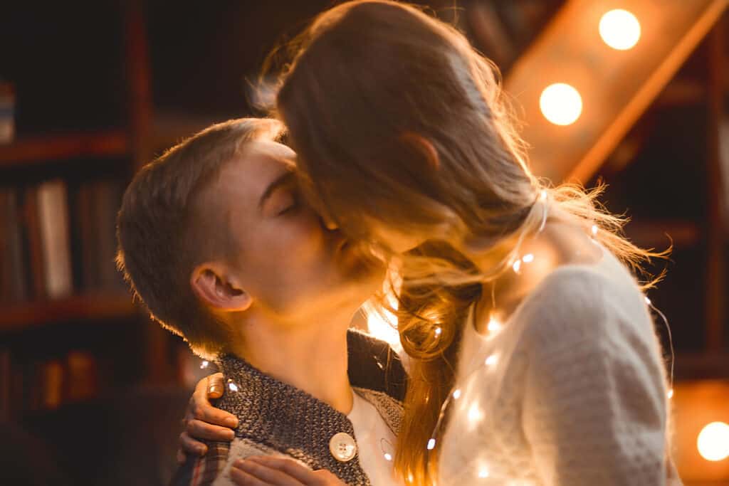 A young couple kisses indoors with bright lights all around them.