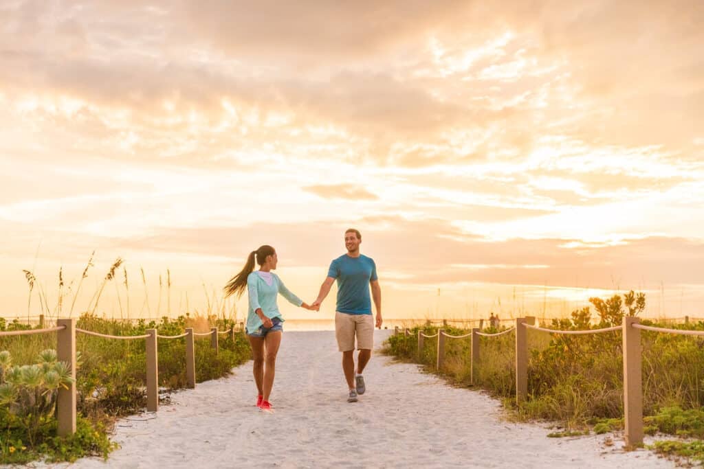 Happy young couple in love walking on romantic beach stroll at sunset. Lovers holding hands on Florida vacation holidays. People walking on summer evening lifestyle.