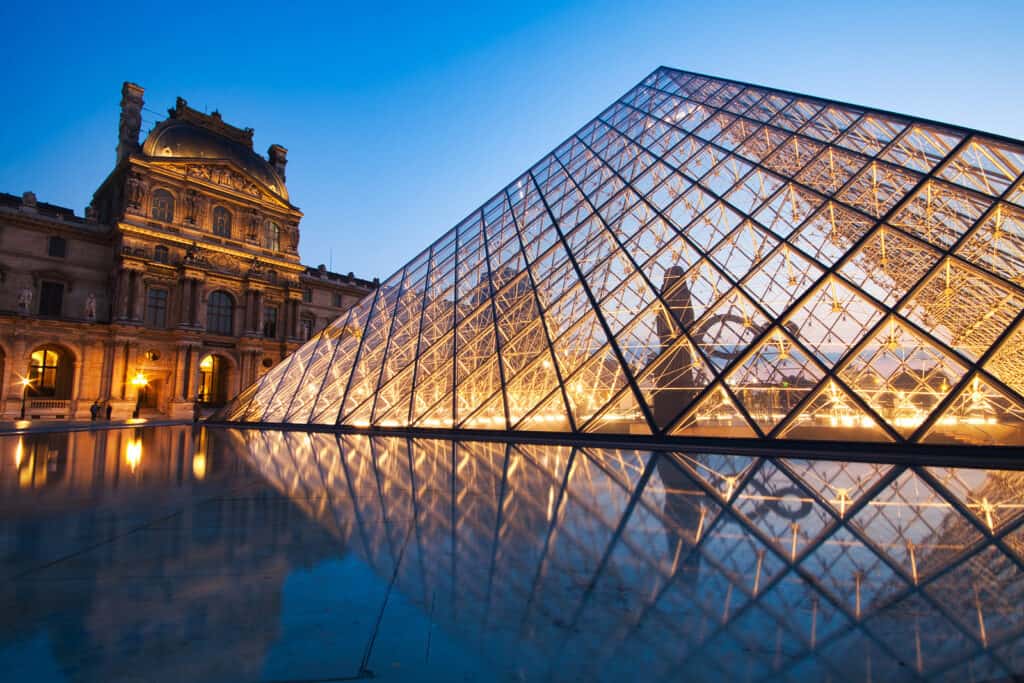 The Louvre is lit up at night under a blue sky.