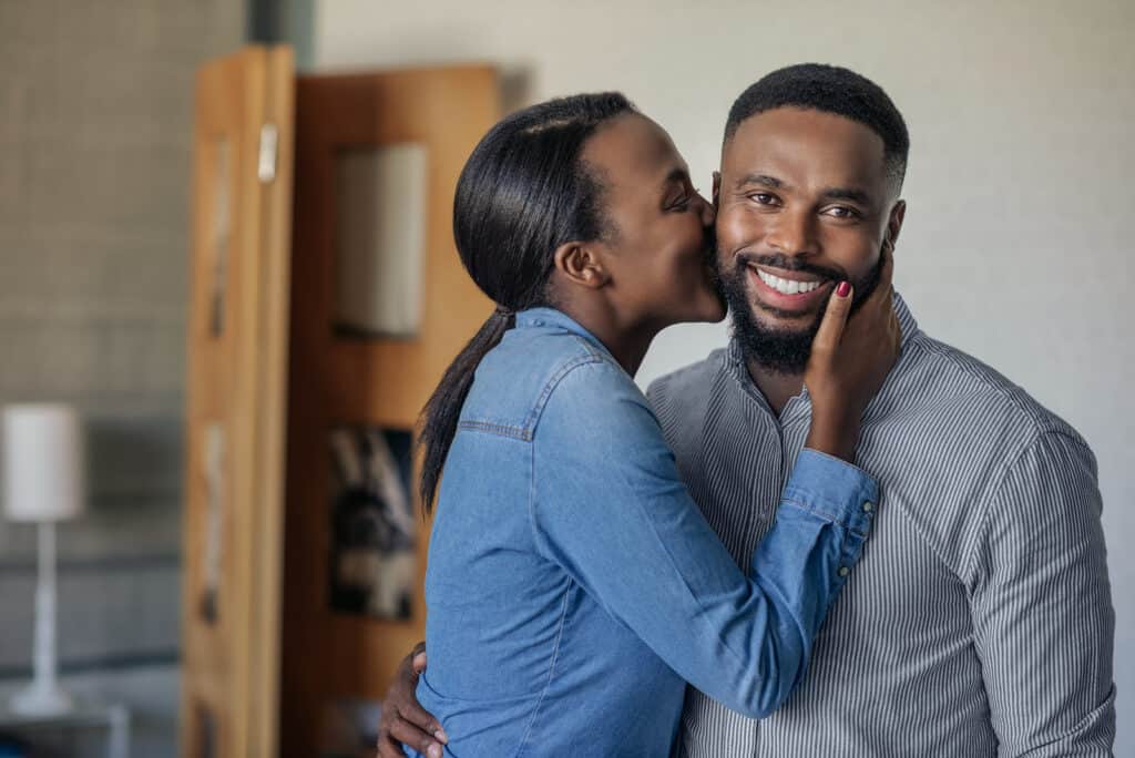 A wife kisses her husband on the cheek while he smiles at the camera.