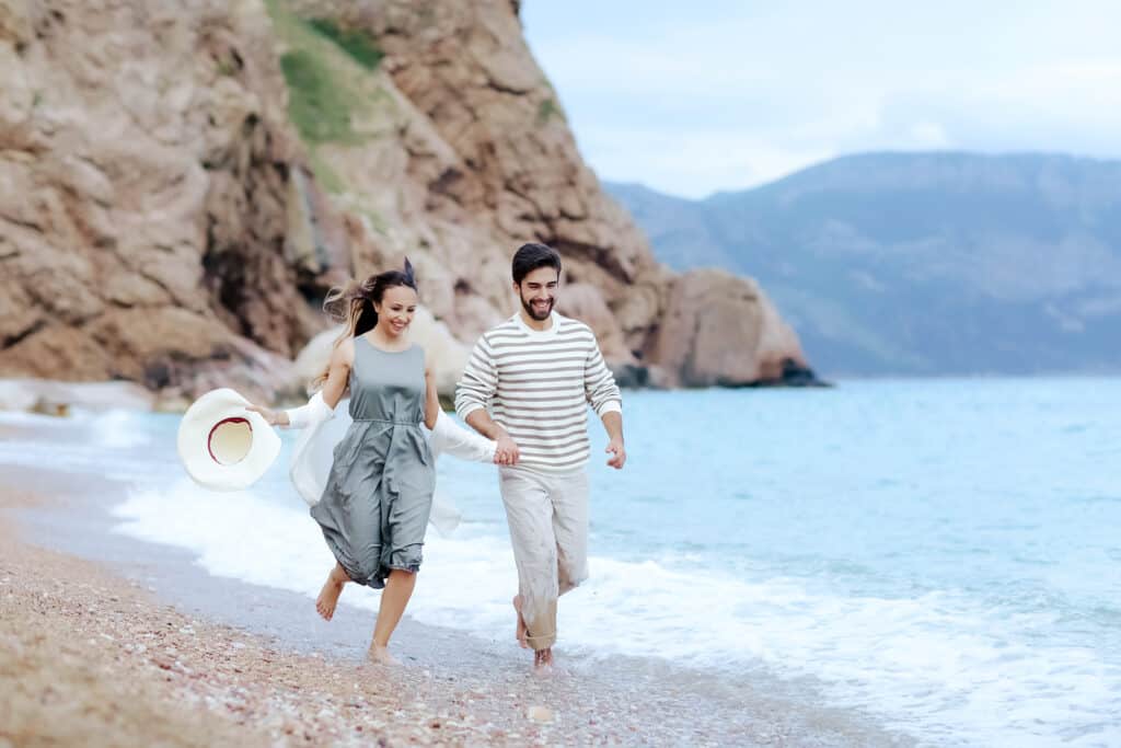 A couple runs through the water on the beach while laughing.