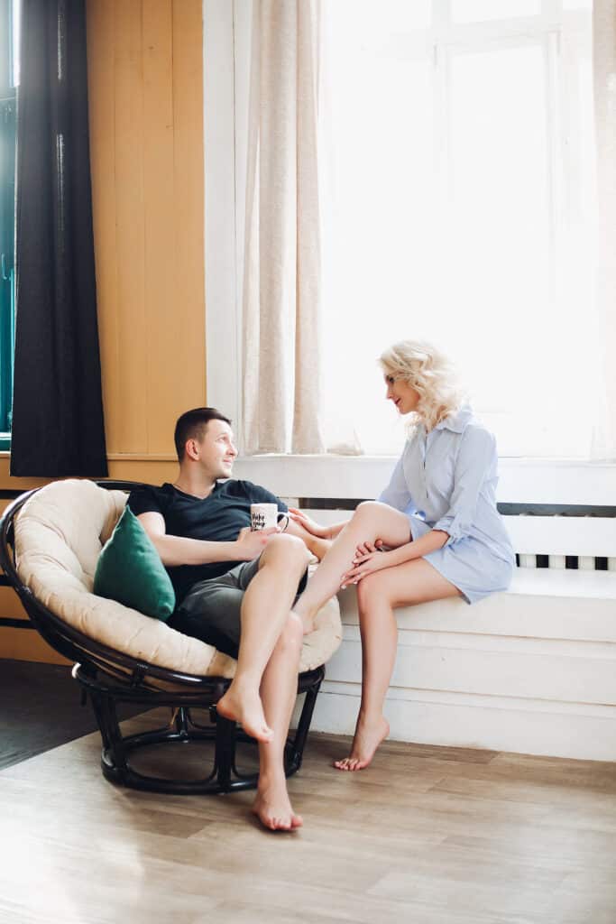 A couple chats while sitting next to a window. The man is holding a coffee mug.