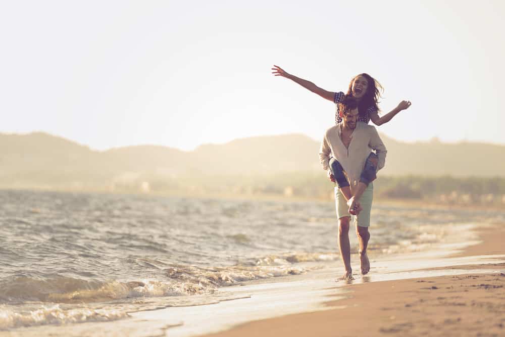 A man holds a woman on his back as they walk along the edge of the beach. 