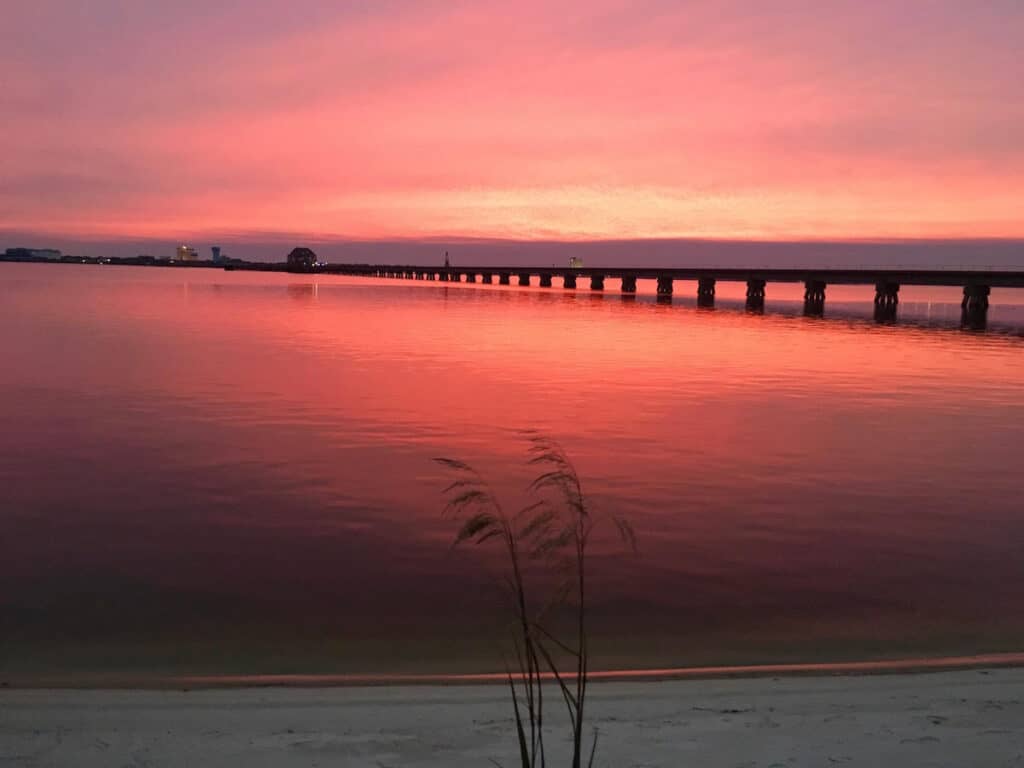 View of the water at the beach with a pier under a blazing pink sunset.