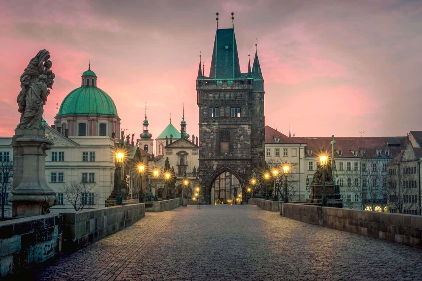 Empty cobblestone walkway leading to a city with green-top roofs. The sky is pink.