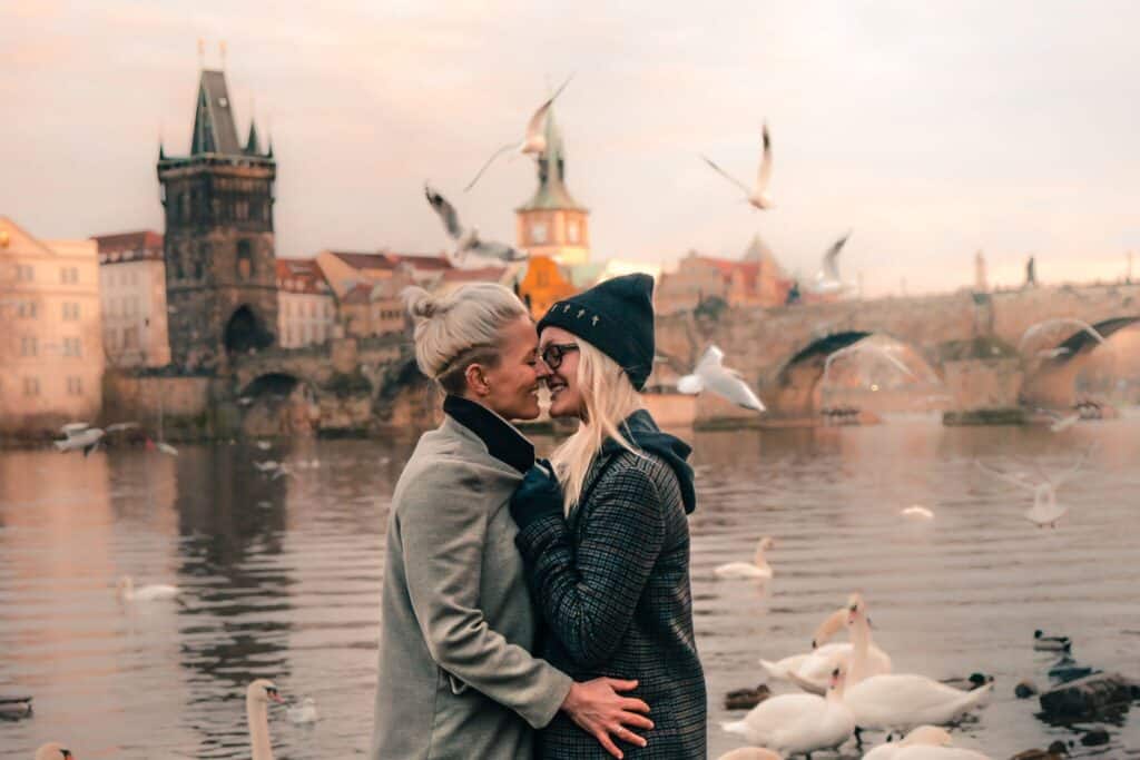 A couple smiles and kisses with birds flying around them next to the water.