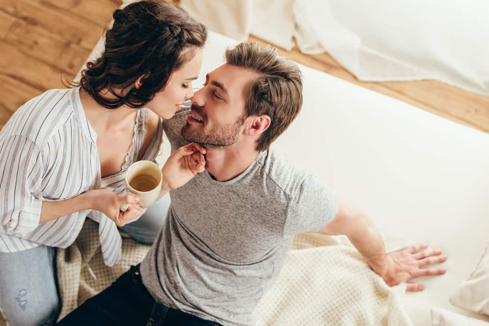 A man and woman are about to kiss while sitting on a bed. The woman holds a cup of coffee.