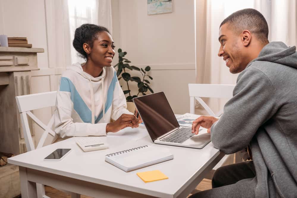 Man and woman chat with each other at a table. The man is on a laptop.