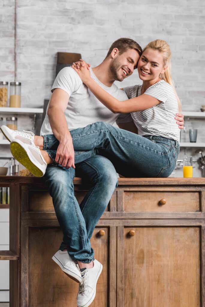 A couple sits on a kitchen island, holding each other and smiling. 