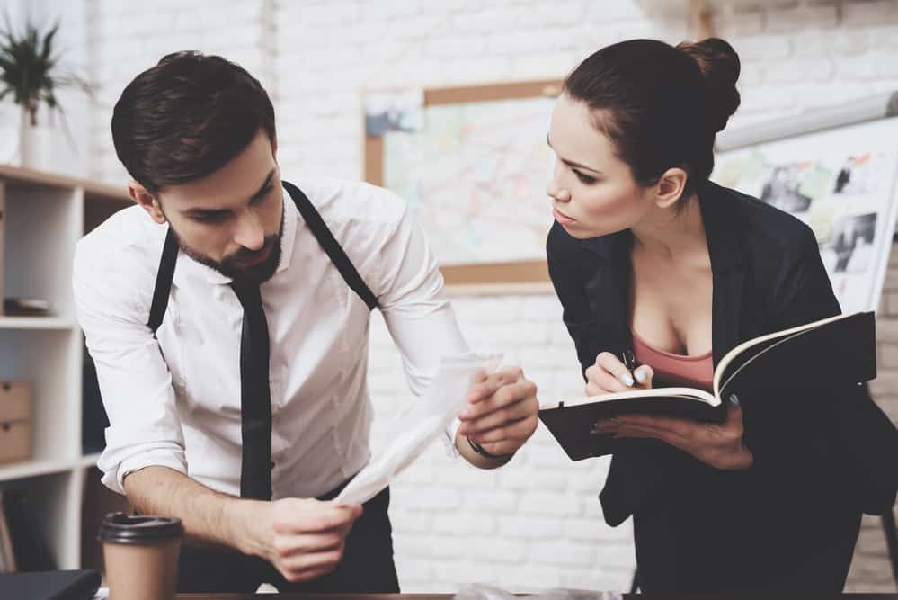 A woman writes in a book while a man holds up a piece of paper.